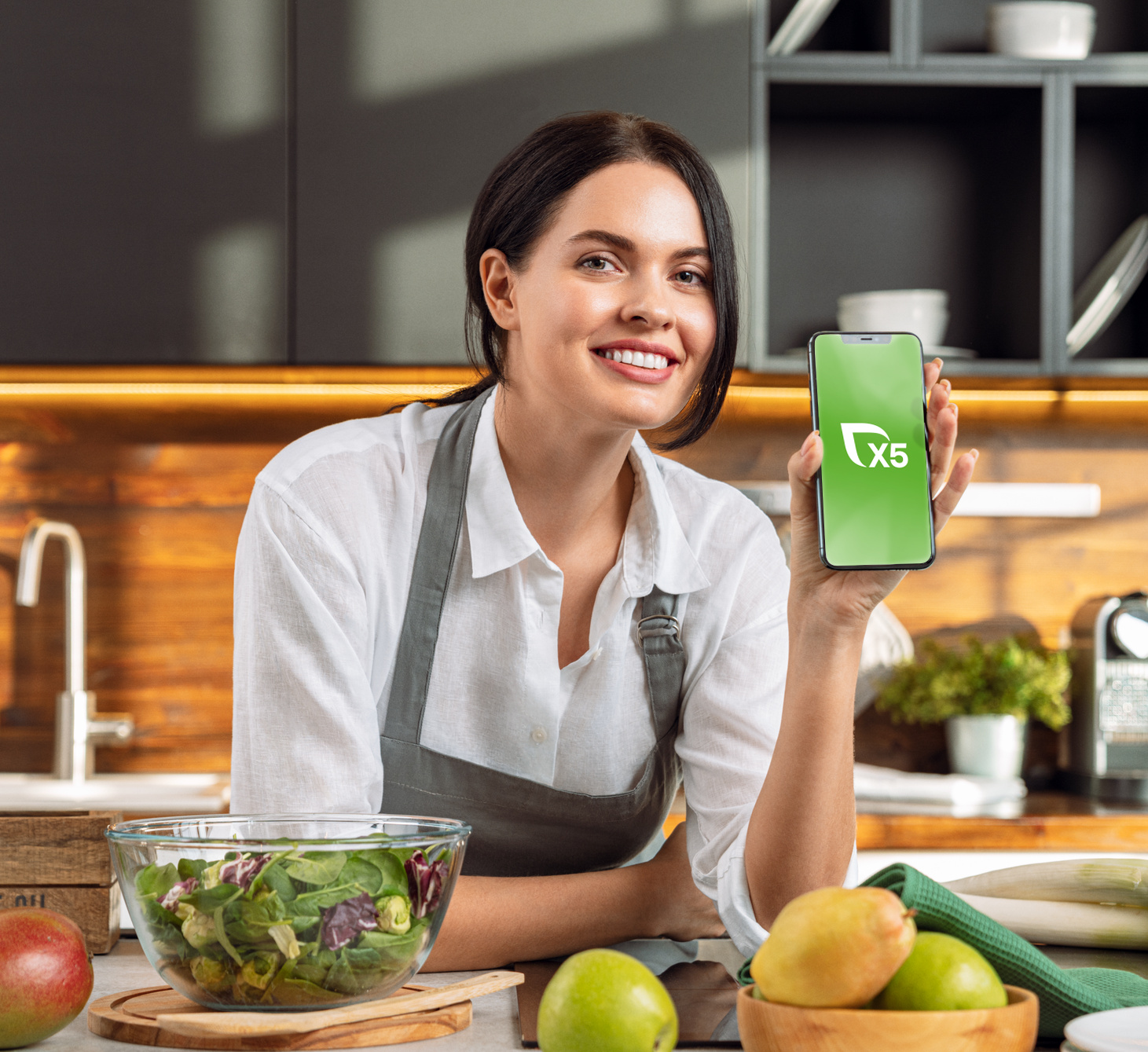 Woman in the kitchen leans on the table with ingredients smiling and shows the phone with the X5 logo
